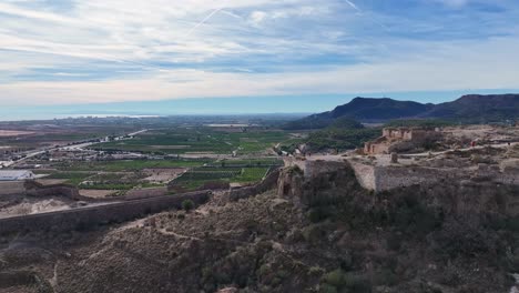 Vuelo-Lateral-Con-Efecto-Paralaje-En-Un-Castillo-Descubriendo-Su-Dimensión-Y-Al-Fondo-Vemos-Campos-De-Cultivos-De-Naranjas-Con-Montañas,-Un-Pueblo-Y-El-Mar,-Hay-Un-Cielo-Azul-Con-Nubes