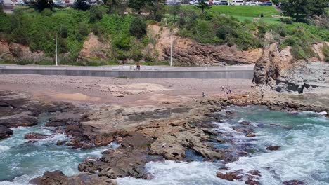 People-and-a-fisherman-on-the-rocks-as-the-surf-washes-in-and-people-walk-on-the-path-beyond