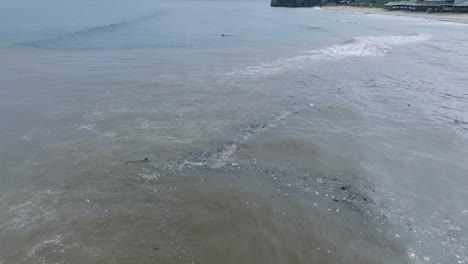 Low-altitude-pan-down-drone-shot-of-polluted-ocean-filled-with-floating-trash-and-debris-and-muddy-sewage-river-runoff-with-surfers-in-the-background-in-Bali-Indonesia