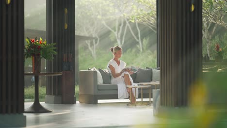 Woman-in-white-dress-sitting-in-luxury-open-resort-lounge-reading-with-morning-light-rays