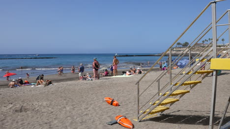 Ein-Sonniger-Strandtag-Mit-Einem-Gelben-Rettungsschwimmerstand-Und-Orangefarbenen-Rettungsringen,-Während-Strandbesucher-Den-Sandstrand-Und-Das-Ruhige-Blaue-Meer-An-Der-Costa-Adeje-Auf-Teneriffa-Genießen