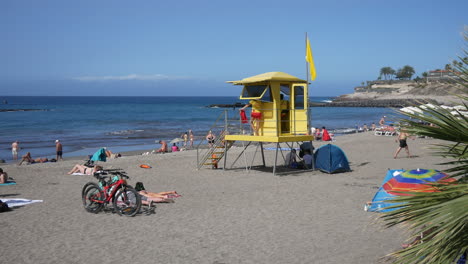 Costa-Adeje,-Tenerife---A-sun-drenched-beach-with-a-yellow-lifeguard-stand,-sunbathers,-palm-trees,-bicycles-and-clear-blue-skies,-exuding-a-relaxed-holiday-vacation-vibe