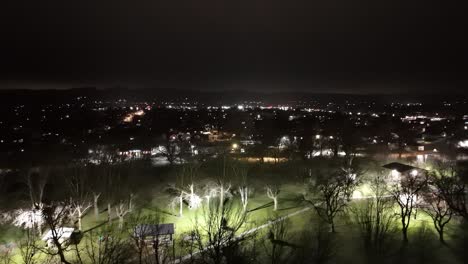 Drone-Vuela-Sobre-El-Parque-De-La-Ciudad-De-Hierba-Verde-En-Una-Pequeña-Ciudad-Por-La-Noche-Mientras-Los-Autos-Pasan-Por-La-Noche