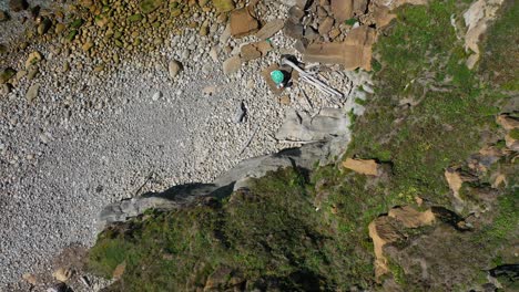 Top-Aufnahme-Einer-Klippe,-An-Der-Eine-Treppe-Entlangführt,-Die-Hinunter-Zum-Steinigen-Strand-Führt,-Wo-Sie-Einige-Sonnenschirme-Und-Die-Wellen-Des-Meeres-Sehen-Können,-Die-Ruhig-Gegen-Die-Felsen-In-Kantabrien,-Spanien,-Schlagen