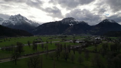 Panorámica-Aérea-A-Través-De-Exuberantes-Campos-Agrícolas-Verdes-Y-árboles-Sin-Hojas-Debajo-De-Las-Montañas-Suizas-En-Walensee