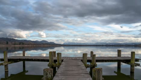 Clouds-reflect-in-serene-water-of-lake-Pfäffikersee-Switzerland-with-empty-wooden-pier-and-stunning-mountains
