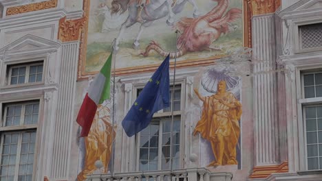 Close-up-clip-of-detailed-murals-at-Palazzo-San-Giorgio,-Genoa,-Italy,-also-showing-Italian-and-EU-flags-flowing-in-breeze