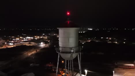 Drohne-Fliegt-Nachts-Im-Nahumlauf-Um-Den-Wassertankturm-Einer-Kleinstadt-Mit-Rotem-Licht-An-Der-Spitze