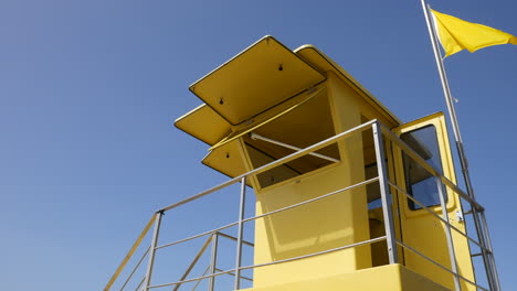 Ein-Gelber-Rettungsschwimmerturm-Vor-Einem-Klaren-Blauen-Himmel,-Eine-Im-Wind-Flatternde-Flagge,-Ein-Symbol-Für-Sicherheit-Und-Wachsamkeit-Im-Sommer-An-Sonnigen-Stränden