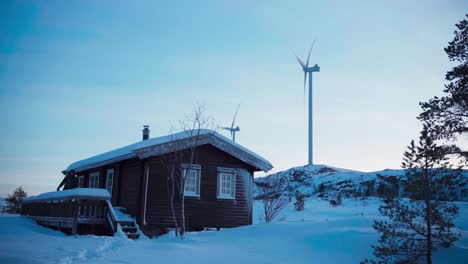 A-Man-Strolling-Away-From-the-Timber-Cabin-Amid-the-Snow-covered-Terrain,-With-Wind-Turbines-Dotting-the-Backdrop-in-Bessaker,-Trondelag-County,-Norway---Static-Shot