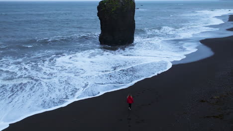 Mujer-Con-Chaqueta-Roja-Caminando-Por-La-Playa-De-Laekjavik-Hacia-El-Océano-En-Islandia