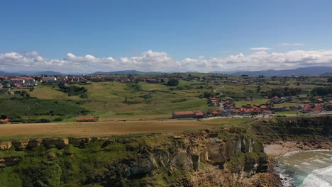 lateral-flight-on-a-cliff-with-a-background-of-mountains-there-are-population-centers-with-scattered-houses-with-green-land-on-the-coast-we-discover-a-beach-with-waves-at-sunset-in-Cantabria-Spain