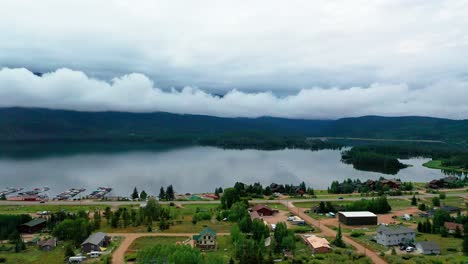 Vista-Aérea-Por-Drones-Del-Hermoso-Embalse-De-Montaña-En-La-Sombra-En-Un-Día-Nublado-De-Verano-En-El-Gran-Lago-Colorado-Con-Automóviles-Conduciendo-Por-La-Carretera-A-Lo-Largo-De-Casas-De-Vacaciones-Pasadas-En-La-Costa-Nublada