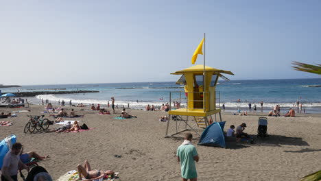 Un-Ajetreado-Día-De-Playa-Con-Un-Puesto-De-Salvavidas-De-Color-Amarillo-Brillante,-Bañistas-Y-Nadadores-En-El-Mar,-Cielos-Azules-Y-Olas-Suaves,-Creando-Una-Vibrante-Atmósfera-De-Vacaciones-En-Costa-Adeje-Tenerife