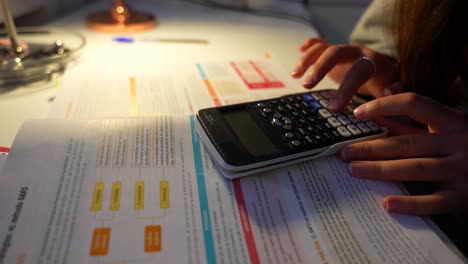 Primer-Plano-De-Delgados-Dedos-Femeninos-Usando-Una-Calculadora-Mientras-Estudia-Tarde-En-La-Noche-Bajo-La-Luz-De-Una-Lámpara-De-Escritorio.