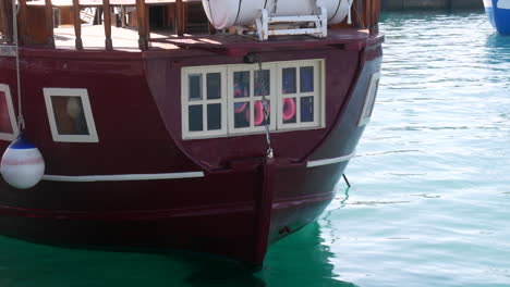 Close-up-of-a-sail-boat-stern-with-quaint-windows-reflecting-life-aboard,-anchored-in-tranquil-turquoise-waters,-hinting-at-maritime-pirate-adventures