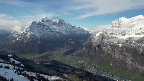 Luftaufnahme-Aus-Der-Vogelperspektive-über-Dem-Dorf-Am-Fuße-Eines-Grünen-Tals-Mit-Schnee--Und-Wolkenbedeckten-Bergen