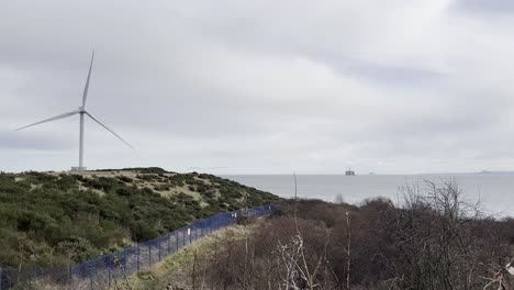 Turbina-Eólica-Girando-Rápidamente-En-Primer-Plano,-Mientras-Que-En-La-Distancia-Se-Ve-Una-Plataforma-Petrolera-En-Alta-Mar-En-El-Mar-Del-Norte