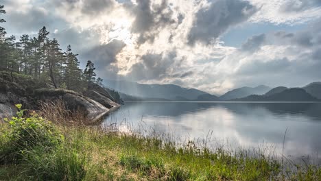 Sonnenstrahlen-Durchdringen-Die-Stürmischen-Wolken,-Die-In-Einem-Zeitraffervideo-über-Dem-See-Und-Einem-Wald-Wirbeln