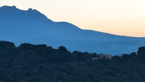 Vuelo-Con-Dron-De-Madrugada-Visualizando-En-166mm-Una-Finca-Con-Una-Casa-Rural-De-Piedra-En-Construcción-Con-El-Fondo-De-Una-Montaña-Creando-Un-Efecto-De-Paralaje-Con-Un-Cielo-Blanquecino-En-Ávila-Invernal