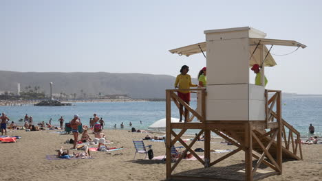 A-bustling-beach-scene-with-a-lifeguard-on-duty,-overseeing-the-safety-of-sunbathers-and-swimmers-against-a-scenic-mountain-backdrop-at-Los-Christianos-in-tenerife