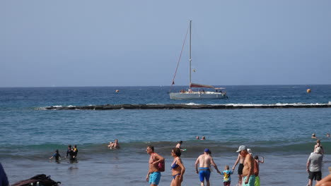 Beach-goers-swim-and-stroll-by-the-water’s-edge-as-a-sailboat-cruises-past-a-rocky-breakwater-under-the-open-sky,-encapsulating-coastal-leisure