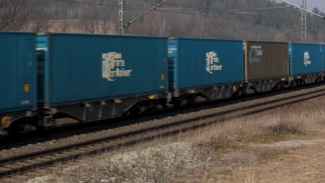Green-locomotive-with-blue-containers-in-motion-on-rural-tracks,-daylight,-blurred-motion-effect