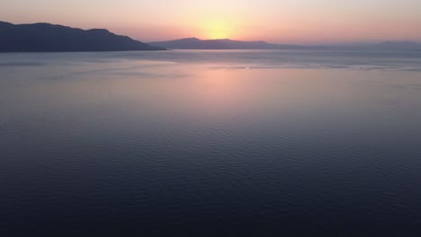 Flight-towards-sunrise-on-the-horizon-over-clear-blue-ocean-water,-aerial-footage,-tilted