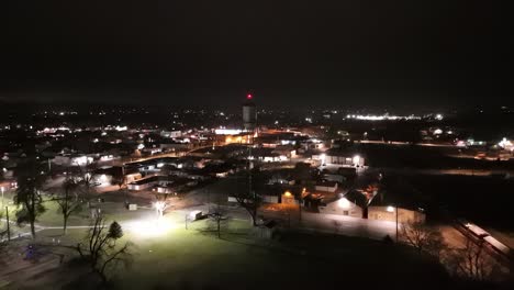 Drone-zooms-into-small-town-water-tank-tower-at-night-near-green-city-park