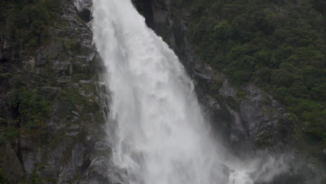 Slow-Motion-pan-down-a-large-waterfall-on-a-cliff-with-a-forest-in-foreground---Milford-Sound-,-New-Zealand