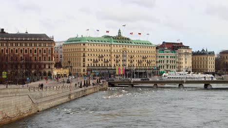 Waterfront-view-of-Grand-Hotel-in-Stockholm-with-flags-waving,-cloudy-sky-above,-and-pedestrians