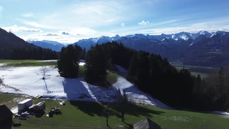 Eine-Drohne-Fliegt-über-Ein-Schneefeld-Und-Enthüllt-Grüne-Grashügel-Und-Einen-Bauernhof-Auf-Den-Bergen-Mit-Herrlichem-Blick-Auf-Schneebedeckte-Berge-An-Einem-Sonnigen-Tag