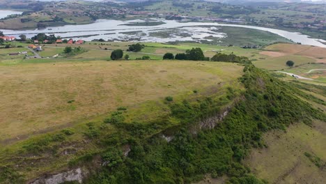 Vuelo-Sobre-Una-Montaña-Llana-Donde-Había-Antiguos-Asentamientos-Viendo-Un-Río-Y-Un-Fondo-De-Montañas-En-Un-Entorno-De-Verdes-Pastos-En-Verano-En-Una-Mañana-Nublada-Cantabria-España