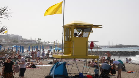 Eine-Lebendige-Strandszene-Mit-Einer-Rettungsschwimmerstation-Mit-Einer-Gelben-Flagge-Auf-Der-Spitze,-Entspannten-Sonnenanbetern,-Sonnenschirmen-Und-Einem-Lebhaften-Ufer-Unter-Dunstigem-Sonnenlicht-An-Der-Costa-Adeje-Auf-Teneriffa