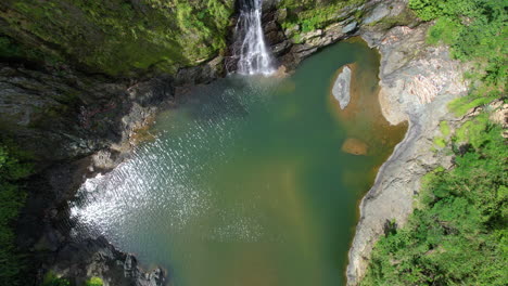 Rising-top-down-drone-shot-of-natural-lake-in-jungle-with-waterfall-at-sunny-day