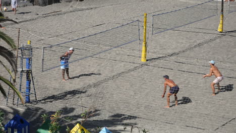 Enérgico-Voleibol-De-Playa-En-Acción,-Con-Jugadores-Masculinos-Saltando-Y-Sacando-En-Una-Cancha-De-Arena,-Enmarcado-Por-Exuberantes-Palmeras-Y-Un-Cielo-Despejado.