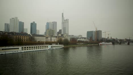 Bewölkter-Blick-Auf-Die-Skyline-Von-Frankfurt-Mit-Main-Und-Vorbeifahrenden-Schiffen,-Kräne-Auf-Baustellen