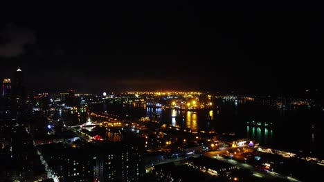 Nighttime-city-skyline-with-twinkling-lights-and-dark-waters-reflecting-urban-glow,-high-angle