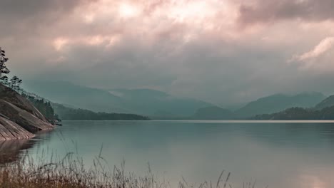 Los-Penetrantes-Rayos-Del-Sol-Brillan-A-Través-De-Las-Nubes-Tormentosas-Que-Giran-Sobre-El-Lago-Con-Bancos-Rocosos-Y-Montañas-En-Un-Vídeo-De-Timelapse