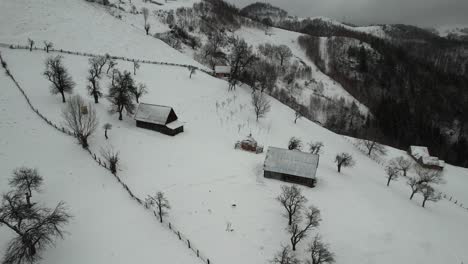 Snowy-cindrel-mountains-with-isolated-cabins,-leafless-trees,-and-winding-trails,-aerial-view