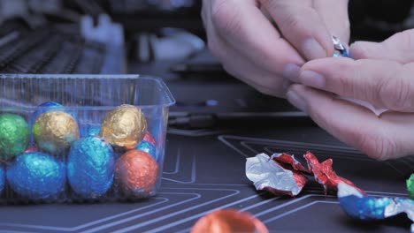 Adult-man-eating-chocolate-Easter-eggs-while-working-on-computer-at-home,-focus-on-hands-taking-colorful-snacks