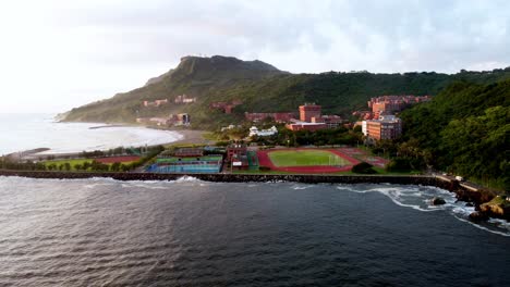 Coastal-town-at-sunrise-with-lush-hills,-sports-fields,-and-calm-ocean,-aerial-view