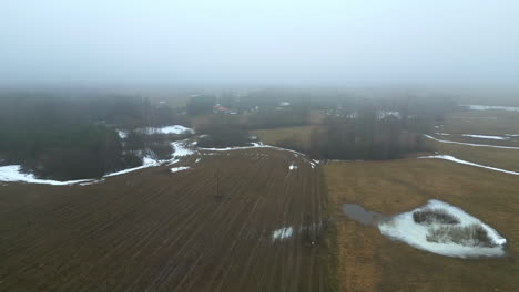 Misty-rural-landscape-in-early-spring-with-melting-snow-patches,-aerial-view