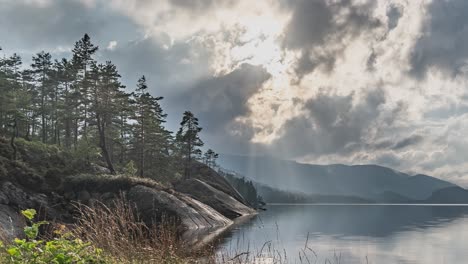 Rays-of-the-sun-pierce-through-the-heavy-stormy-clouds-above-the-still-lake-with-rocky-banks-covered-with-pine-forest