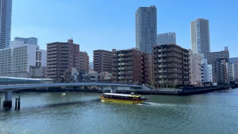 Gelbes-Boot,-Das-Auf-Dem-Fluss-In-Städtischer-Umgebung-Kreuzt,-Stadtgebäude-Im-Hintergrund,-Klarer-Himmel