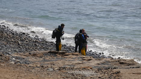 Scuba-divers-with-diving-equipment-ready-for-an-ocean-adventure,-standing-on-a-rocky-beach-as-waves-gently-roll-in