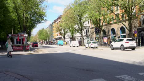 City-life-in-central-Stockholm-with-pedestrians-crossing-Sveavägen-street,-sunlit-buildings-and-lush-trees,-daytime