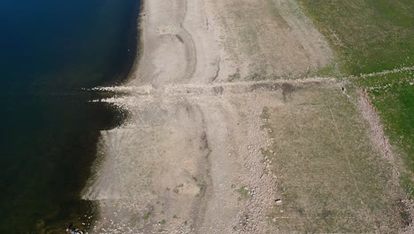 Vuelo-Con-Dron-Por-La-Orilla-De-Un-Embalse-Donde-Vemos-El-Agua,-La-Tierra-Blanquecina-Con-Restos-De-Piedras-Formando-Estructuras-Con-Un-Camino-Sumergido-Y-Un-Terreno-Con-Plantas-Verdes-Invernando-En-España