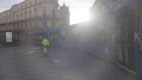 Street-Cleaner-Walking-in-Montpellier,-Blue-Tramway-Stopped,-Backdropped-by-Beautiful-Sunset-and-Historic-Buildings
