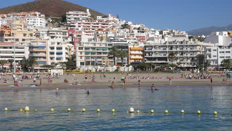 Vibrant-beach-life-with-sunbathers-and-swimmers-against-a-bustling-city-hotels-backdrop,-embodying-leisure-and-urban-coastal-living-at-Los-Christianos-beach-in-Tenerife